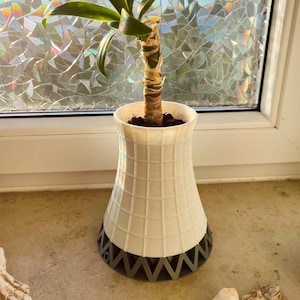Könnte beinhalten: Ein weiß-grauer dekorativer Blumentopf mit einem Gittermuster, das an einen Kühlturm erinnert. Der Topf enthält eine kleine Pflanze mit grünen Blättern und einem braunen Stiel. Der Topf steht auf einer Fensterbank.