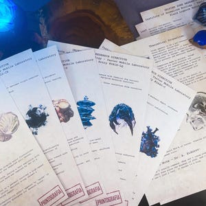 Pode incluir: Uma coleção de documentos impressos com diagramas científicos e texto, apresentando várias ilustrações abstratas em azul, preto e castanho. Os papéis estão dispostos numa superfície com uma fonte de luz azul no fundo, criando uma atmosfera misteriosa.