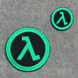 Puede incluir: Dos parches circulares con un s&iacute;mbolo lambda verde sobre un fondo negro, rodeados por un borde verde. Los parches son de diferentes tama&ntilde;os, con el m&aacute;s grande en primer plano. Estos parches son probablemente para ropa o accesorios.