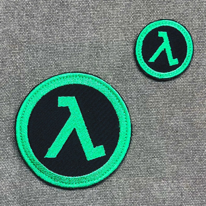 Puede incluir: Dos parches circulares con un s&iacute;mbolo lambda verde sobre un fondo negro, rodeados por un borde verde. Los parches son de diferentes tama&ntilde;os, con el m&aacute;s grande en primer plano. Estos parches son probablemente para ropa o accesorios.