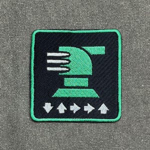 May include: Embroidered patch with a black background and a green border. The patch features a green design of a stylized cannon with white horizontal lines and white arrows pointing up and down.