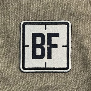 Könnte beinhalten: Quadratischer Aufnäher mit weißem Hintergrund und schwarzem Rand. Die Buchstaben "BF" sind in fetter schwarzer Schrift in der Mitte. Der Aufnäher hat fadenkreuzartige Markierungen oben, unten und an den Seiten. Der Hintergrund ist strukturiert und neutral gefärbt.