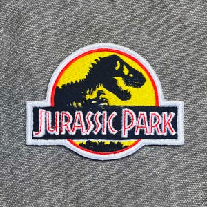 May include: Embroidered patch with the "Jurassic Park" logo. The design features a black Tyrannosaurus Rex silhouette on a yellow background, a red border, and the words "JURASSIC PARK" in red letters on a white background.