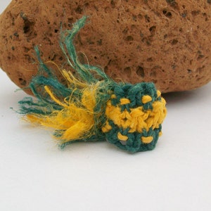 May include: A small, handmade crochet item in shades of green and yellow, with a textured appearance. The item has a rounded shape with a fringe of loose yarn. A rough, brown stone is in the background.
