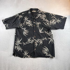 May include: Black button-down shirt with short sleeves and a bamboo leaf pattern in light beige. The shirt has a collared neckline and a pocket on the left chest. The fabric appears to be lightweight.