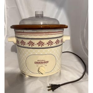 May include: Cream-colored Rival Crock-Pot slow cooker with a brown rim and clear lid. The cooker has a floral pattern and the text "RIVAL CROCK-POT" on the front. It features a dial for low, high, and off settings, and a brown power cord.