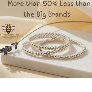 May include: Three silver beaded bracelets are stacked on a white surface, with a sprig of lavender and text that reads "More than 50% Less than the Big Brands." The Charmed Bee logo is in the upper left corner.
