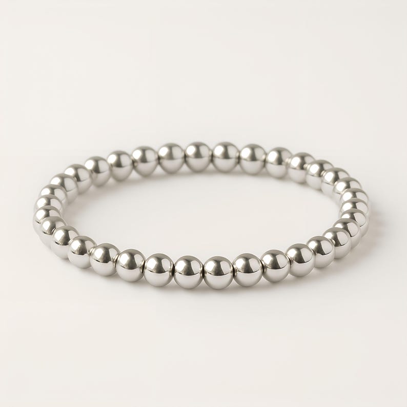 K&ouml;nnte beinhalten: Ein silbernes Perlenarmband. Das Armband besteht aus kleinen, runden, gl&auml;nzenden Silberperlen, die zu einer kreisf&ouml;rmigen Form aufgereiht sind. Die Perlen sind gleichm&auml;&szlig;ig verteilt und reflektieren das Licht, wodurch ein heller, metallischer Glanz entsteht.