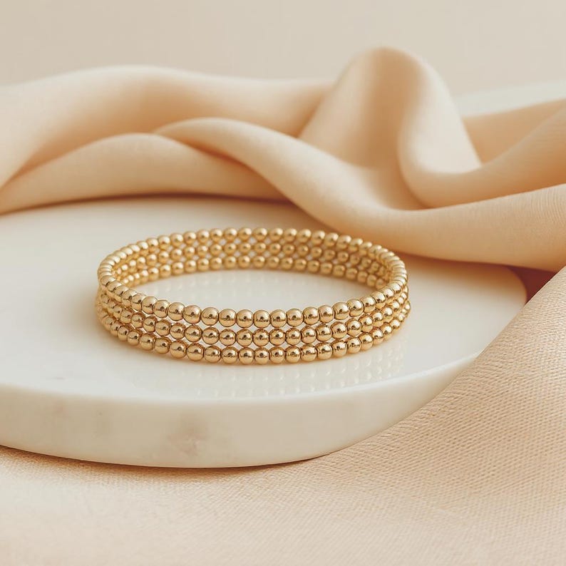 K&ouml;nnte beinhalten: Ein goldfarbenes Armband mit drei Reihen kleiner, runder Perlen. Das Armband liegt auf einer wei&szlig;en, runden Oberfl&auml;che, im Hintergrund ein weicher, pfirsichfarbener Stoff. Ein schlichtes, elegantes Schmuckst&uuml;ck.