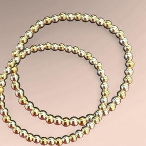K&ouml;nnte beinhalten: Zwei Armb&auml;nder mit Perlen, eines goldfarben und eines silberfarben, vor einem sanften rosa Hintergrund. Jedes Armband besteht aus kleinen, runden Perlen, die aufgef&auml;delt sind. Die Perlen reflektieren das Licht und erzeugen einen subtilen Schimmer.