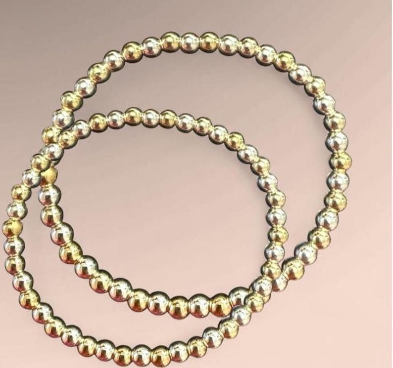 K&ouml;nnte beinhalten: Zwei Armb&auml;nder mit Perlen, eines goldfarben und eines silberfarben, vor einem sanften rosa Hintergrund. Jedes Armband besteht aus kleinen, runden Perlen, die aufgef&auml;delt sind. Die Perlen reflektieren das Licht und erzeugen einen subtilen Schimmer.