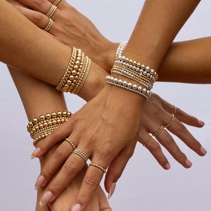 K&ouml;nnte beinhalten: Nahaufnahme mehrerer H&auml;nde, die mit gold- und silberfarbenen Perlenarmb&auml;ndern und Ringen geschm&uuml;ckt sind. Der Schmuck weist Perlen in verschiedenen Gr&ouml;&szlig;en auf, wodurch ein geschichteter und strukturierter Look entsteht. Die Ringe sind zart und erg&auml;nzen die Armb&auml;nder. Die Gesamt&auml;sthetik ist elegant und stilvoll.