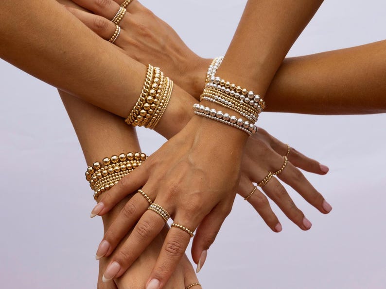 K&ouml;nnte beinhalten: Nahaufnahme mehrerer H&auml;nde, die mit gold- und silberfarbenen Perlenarmb&auml;ndern und Ringen geschm&uuml;ckt sind. Der Schmuck weist Perlen in verschiedenen Gr&ouml;&szlig;en auf, wodurch ein geschichteter und strukturierter Look entsteht. Die Ringe sind zart und erg&auml;nzen die Armb&auml;nder. Die Gesamt&auml;sthetik ist elegant und stilvoll.