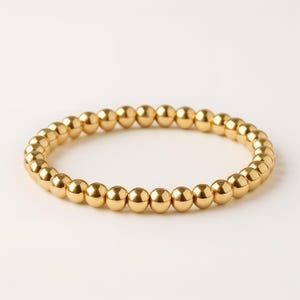 K&ouml;nnte beinhalten: Ein goldfarbenes Perlenarmband. Das Armband besteht aus kleinen, runden, goldfarbenen Perlen, die zu einer kreisf&ouml;rmigen Form aufgereiht sind. Die Perlen haben eine gl&auml;nzende, reflektierende Oberfl&auml;che. Das Armband wird auf wei&szlig;em Hintergrund gezeigt.