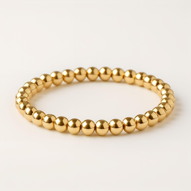 K&ouml;nnte beinhalten: Ein goldfarbenes Perlenarmband. Das Armband besteht aus kleinen, runden, goldfarbenen Perlen, die zu einer kreisf&ouml;rmigen Form aufgereiht sind. Die Perlen haben eine gl&auml;nzende, reflektierende Oberfl&auml;che. Das Armband wird auf wei&szlig;em Hintergrund gezeigt.