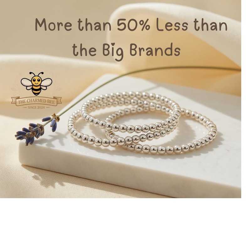 K&ouml;nnte beinhalten: Drei silberne Perlenarmb&auml;nder sind auf einer wei&szlig;en Oberfl&auml;che gestapelt, mit einem Lavendelzweig und dem Text "More than 50% Less than the Big Brands". Das Logo von The Charmed Bee befindet sich in der oberen linken Ecke.