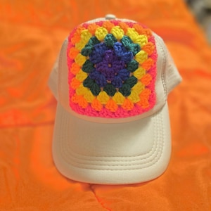 May include: A cream-colored baseball cap featuring a square, multi-colored crochet patch on the front panel. The patch is composed of concentric layers of yarn in bright shades of pink, orange, yellow, dark green, and blue in a square pattern.
