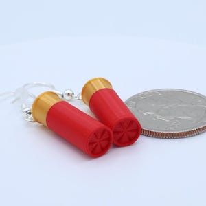 May include: A pair of red and gold shotgun shell earrings with silver hooks. The earrings are next to a quarter coin for size comparison. The earrings have a detailed design on the bottom.