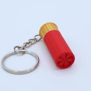 May include: A red and gold shotgun shell keychain with a silver chain and keyring. The keychain is a novelty item, likely made of plastic or resin, and is designed to resemble a real shotgun shell.