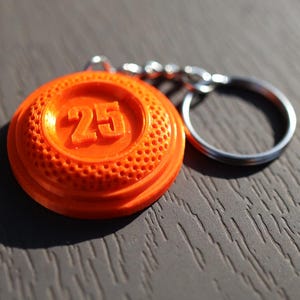 May include: Orange keychain with a round, textured disc featuring the number "25" in raised lettering. The keychain is attached to a silver metal ring.