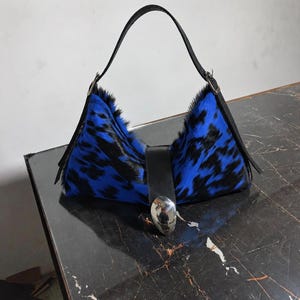 May include: A hobo-style handbag featuring a vibrant blue and black animal print fur texture. The bag includes a black leather strap, a central black leather vertical tab, and a large, polished silver-tone metallic oval accent at the bottom.