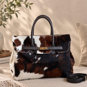 May include: A handbag featuring a cowhide pattern with patches of brown, black, and white fur. The bag has dark brown leather handles, a horizontal strap across the front with a gold-toned metal clasp, and a detachable black leather shoulder strap.