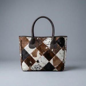 May include: A tote bag featuring a patchwork design of cowhide squares in shades of brown, black, and white. The bag has two dark brown leather handles attached to the top edge and a structured rectangular shape with dark brown leather trim.