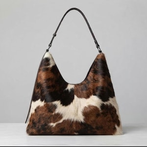 Könnte beinhalten: Eine Schultertasche im Hobo-Stil mit einem strukturierten Rindslederexterieur, das ein gesprenkeltes Muster aus braunen, schwarzen und weißen Flecken aufweist. Die Tasche hat einen einzelnen dünnen schwarzen Lederriemen mit Metallbeschlägen.