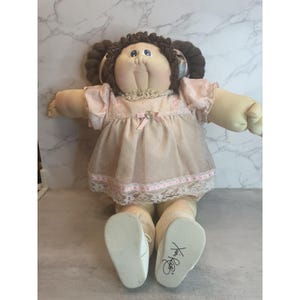 May include: A vintage Cabbage Patch Kid doll with brown yarn pigtails. The doll is dressed in a pink dress with lace trim and a bow. The doll's feet are wearing white shoes, and the soles are signed.