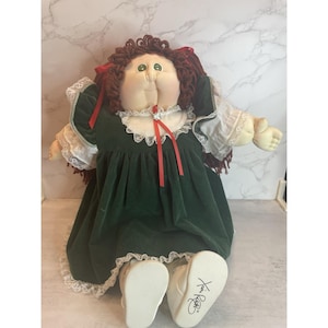 Peut inclure: Poupée vintage avec un visage doux et des cheveux bruns en fil. La poupée porte une robe en velours vert avec une garniture en dentelle blanche et des rubans rouges. Les chaussures blanches de la poupée ont une signature sur la semelle.