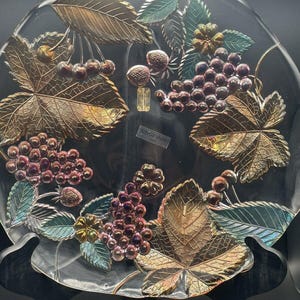 Puede incluir: Plato decorativo de cristal transparente con hojas doradas en relieve, uvas moradas y cerezas rojas. El plato tiene un borde festoneado y está sostenido por un soporte negro. El texto "savor vivre" es visible.