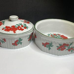 May include: A white ceramic baking dish with a lid, both featuring a red and green floral pattern. The dish has a fluted edge and the lid has a knob.