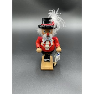 Könnte beinhalten: Eine Holz-Nussknacker-Figur in einem roten Mantel, einer schwarzen Hose und einem schwarzen Zylinder mit weißen Federn. Die Figur hat einen weißen Bart, blaue Augen und hält eine kleine Uhr. Sie steht auf einem Holzsockel.