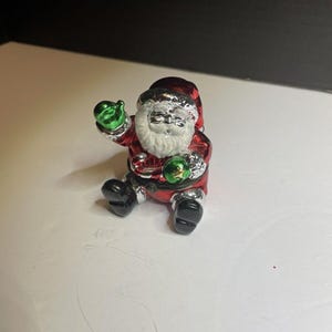 May include: A small, silver-colored figurine of Santa Claus sitting with a green gift in his hand. He is wearing a red suit with white trim and a white beard.