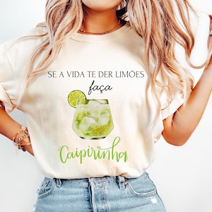 Könnte beinhalten: Cremefarbenes T-Shirt mit dem Text "SE A VIDA TE DER LIMÕES faça Caipirinha" in Schwarz und Grün. Das Design beinhaltet eine Aquarell-Illustration eines Caipirinha-Cocktails mit Limettenscheiben und Eis.