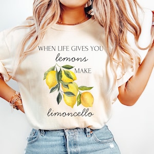 Puede incluir: Camiseta color crema con el texto "WHEN LIFE GIVES YOU lemons MAKE limoncello" sobre una ilustración de acuarela de limones y hojas verdes. La camiseta es de estilo casual.
