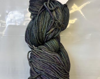 May include: A skein of dark gray yarn with subtle green and purple highlights. The yarn is labeled "863 Zarzamora Rios" and appears to be a worsted weight. The yarn is tightly wound and ready for knitting or crocheting projects.