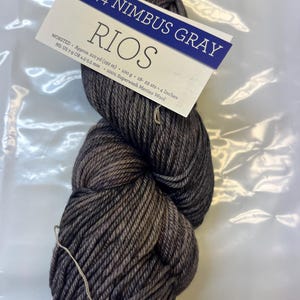 Könnte beinhalten: Ein Knäuel Rios-Garn in der Farbe Nimbus Grau. Das Garn ist ein Wollgarn und besteht aus 100% supergewaschener Merinowolle. Das Knäuel ist 210 Yards (192 Meter) lang und wiegt 100 Gramm. Die Nadelstärke beträgt US 7-9 oder 4,5-5,5 mm. 18-22 Maschen entsprechen 4 Zoll.