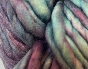 Puede incluir: Primer plano de una lana suave y gruesa en tonos de morado, turquesa y verde. La lana está retorcida y apilada, creando un aspecto texturizado y colorido.
