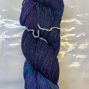 May include: A skein of deep blue yarn with purple and teal colour variations. The yarn is wound into a ball shape and is ready for knitting or crocheting.
