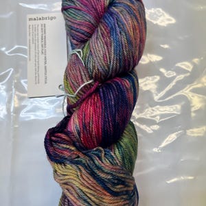 Puede incluir: Una madeja de hilo Malabrigo en una vibrante mezcla de colores, incluyendo rosa, morado, azul y verde. El hilo est&aacute; bien enrollado y parece suave. Una etiqueta blanca con el nombre de la marca e instrucciones de cuidado es visible.