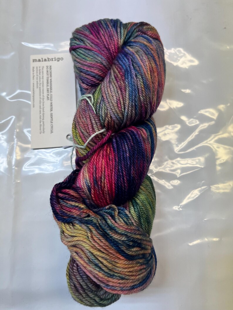 Puede incluir: Una madeja de hilo Malabrigo en una vibrante mezcla de colores, incluyendo rosa, morado, azul y verde. El hilo est&aacute; bien enrollado y parece suave. Una etiqueta blanca con el nombre de la marca e instrucciones de cuidado es visible.