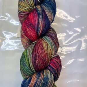 Puede incluir: Un ovillo de lana colorida con una mezcla de tonos vibrantes. La lana presenta una mezcla de azules, verdes, rojos y amarillos, creando un efecto degradado visualmente atractivo. La lana est&aacute; bien enrollada, lo que sugiere que est&aacute; lista para proyectos de tejido o ganchillo.