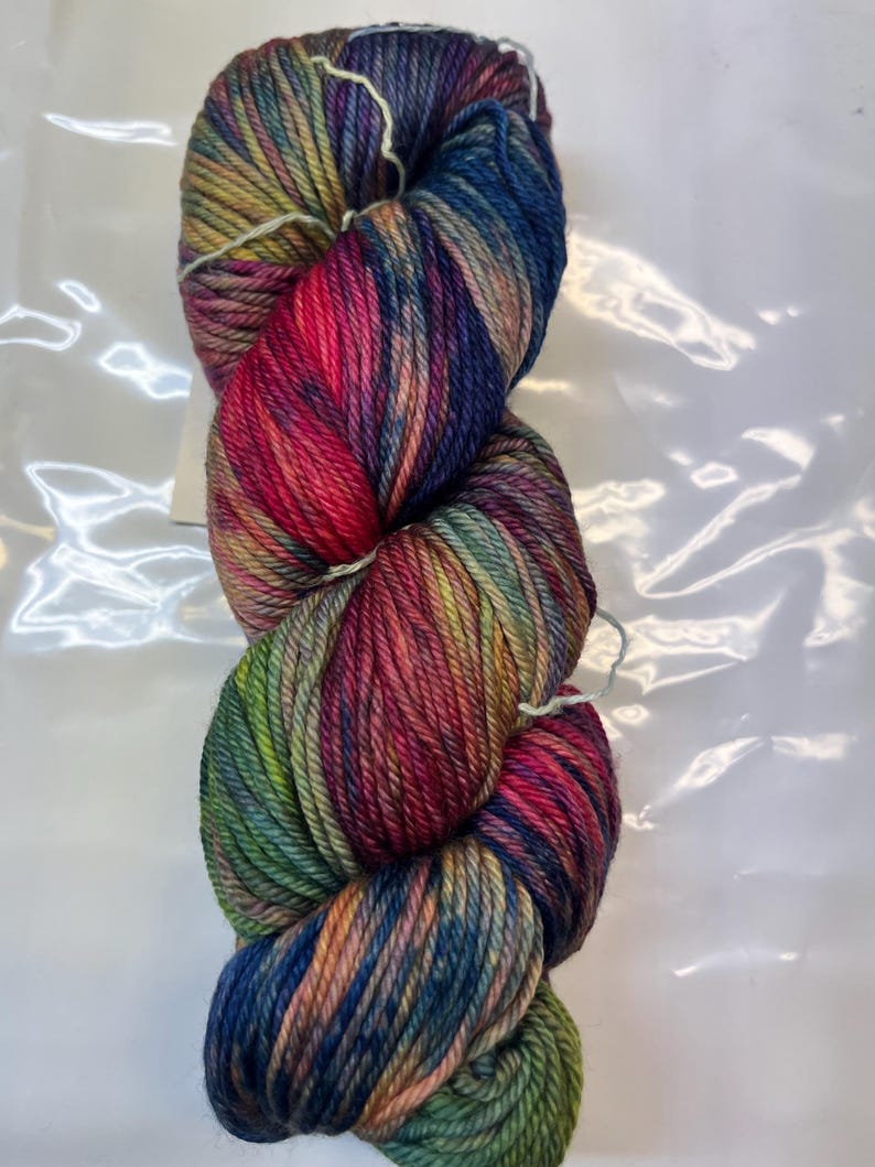 Puede incluir: Un ovillo de lana colorida con una mezcla de tonos vibrantes. La lana presenta una mezcla de azules, verdes, rojos y amarillos, creando un efecto degradado visualmente atractivo. La lana est&aacute; bien enrollada, lo que sugiere que est&aacute; lista para proyectos de tejido o ganchillo.