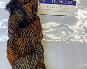 May include: A skein of yarn in shades of brown, green, purple, and black. The yarn is labeled "862 Piedras Mecha".