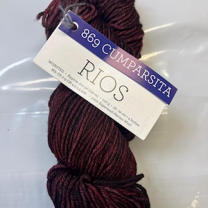 May include: A skein of dark red yarn labelled "869 Cumparsita" with the brand name "RIOS" and details about the yarn: "WORSTED Approx. 210 yd (192 m) 100 g. 18-22 sts = 4 Inches NS: US 7-9 OR 4-5-5-5 mm 100% Superwash Merino Wool"