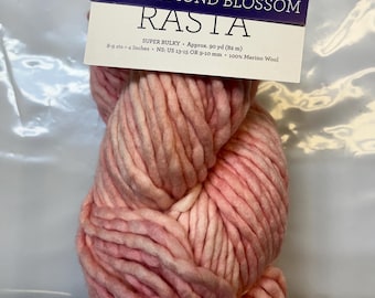 Puede incluir: Un gran ovillo de lana Rasta súper gruesa rosa y blanca. La etiqueta dice "703 Almond Blossom" e incluye detalles sobre el peso, la longitud (aproximadamente 82 metros) y la tensión (8-9 puntos por 10 cm). Está hecha de 100% lana merino.