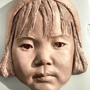 May include: A terracotta-colored clay wall plaque featuring a sculpted face of a child with a bob haircut and bangs. The facial features include almond-shaped eyes, a small nose, and a neutral expression. The texture shows visible tool marks.