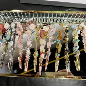 Op de afbeelding: Een verzameling decoratieve clips met diverse versieringen, waaronder kralen, kristallen en bloemaccenten. De clips worden gepresenteerd op een spiegeltray, met een scala aan kleuren en texturen, met goud-, zilver- en koperkleurige metalen clips.