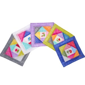 May include: A collection of colorful, square fabric coasters. Each coaster features a patchwork design with geometric shapes in various colors, including pink, blue, green, and orange. Small embroidered floral details are present on each coaster.